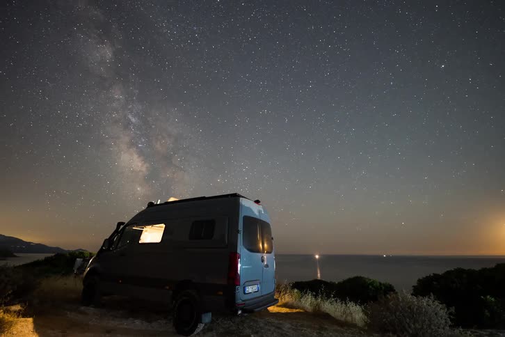 A van sitting still by the coastline as the setting sun disappears turning evening into night with the bright night sky illuminated by stars