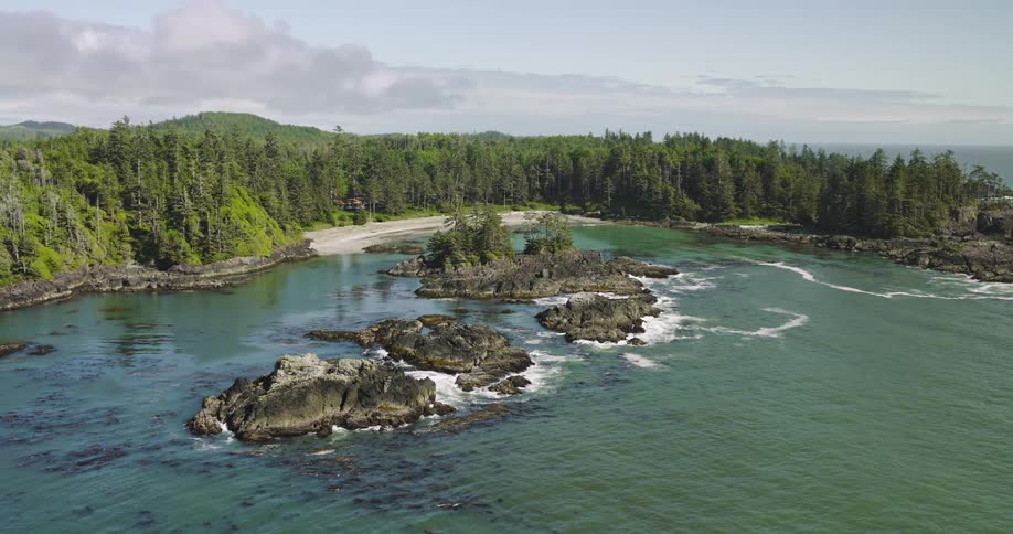 A majestic aerial view showcases a rugged Pacific coastline, revealing a secluded cove with turquoise water, rocky islands, and a dense evergreen forest