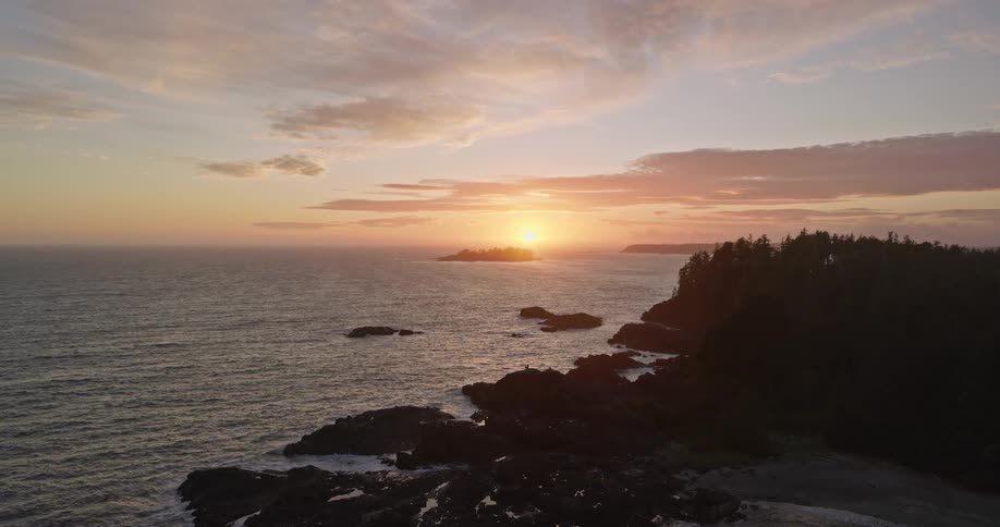 A serene aerial shot captures the golden sunset over a rugged Pacific coastline with rocky outcrops and a dense forest
