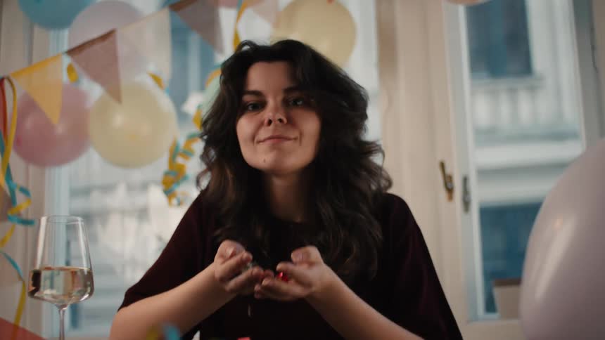 Joyful young woman with dark hair blows a handful of colorful confetti towards the camera in slow motion, surrounded by party decorations