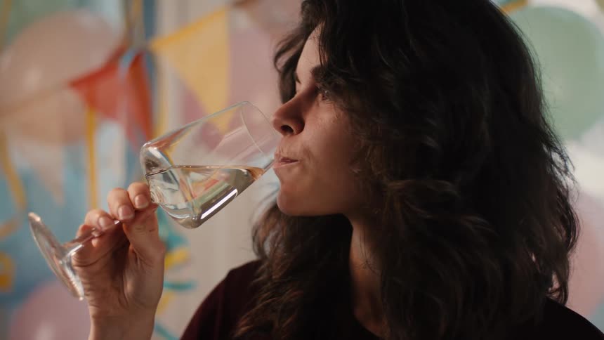 A beautiful woman with dark curly hair smiles contentedly as she sips white wine at a birthday party in slow motion