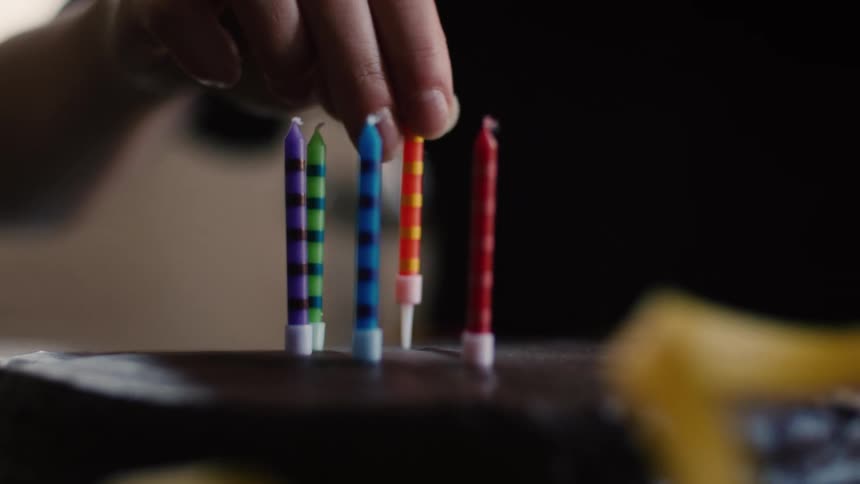A hand carefully places colorful striped birthday candles onto a chocolate cake in slow motion