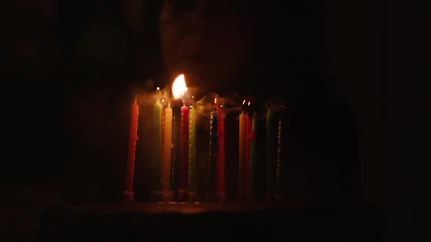 A person in a dark room blows out colorful birthday candles on a cake in slow motion, making a wish