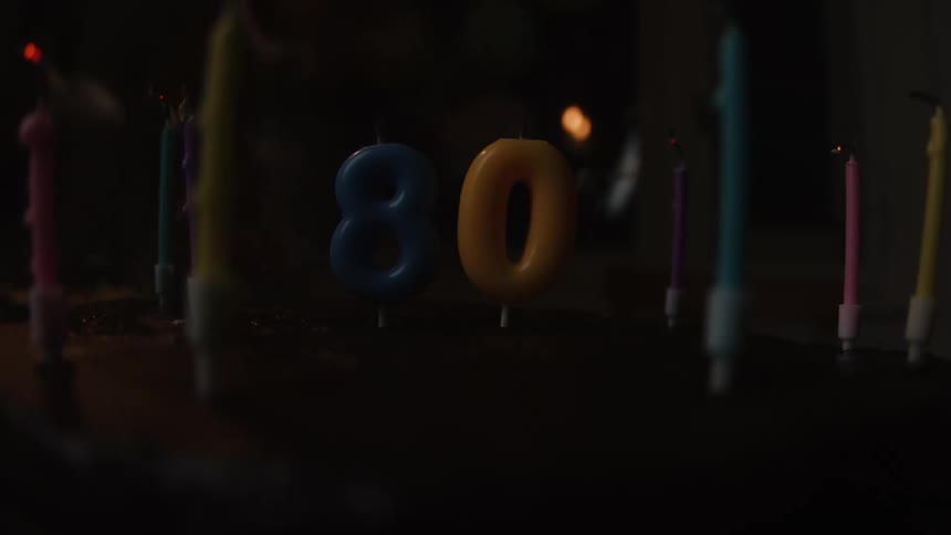 Candles for an 80th birthday are blown out on a cake in a dark room