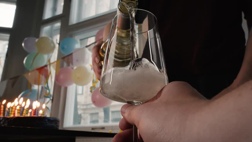 A first-person view of sparkling champagne being poured into a glass in slow motion during a birthday party with a cake and lit candles in the background