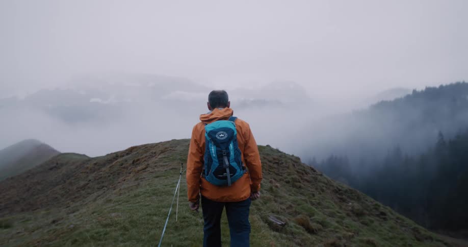 A lone hiker in an orange jacket walks along a grassy mountain crest into the thick fog