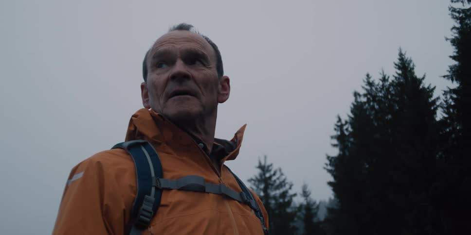 A thoughtful older man in an orange jacket with a backpack pauses to look around while hiking in a foggy forest