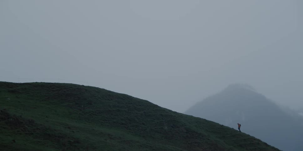 A solitary hiker with a backpack slowly ascends a vast, grassy mountainside on a moody and foggy day