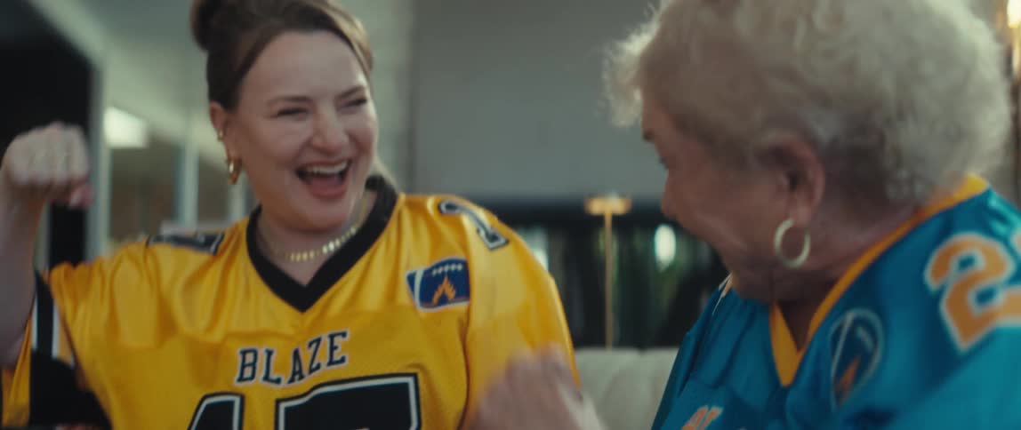A young woman and an elderly woman in football jerseys laugh and fist bump in slow motion while watching a game on a tablet