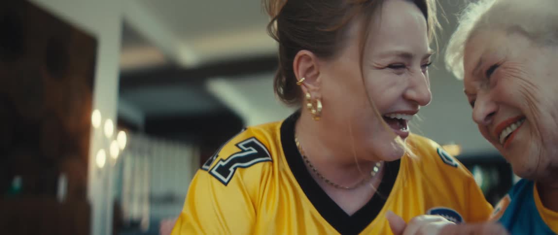 A young woman and her elderly mother share a joyful moment, playfully arm wrestling and laughing on the couch in rival football jerseys