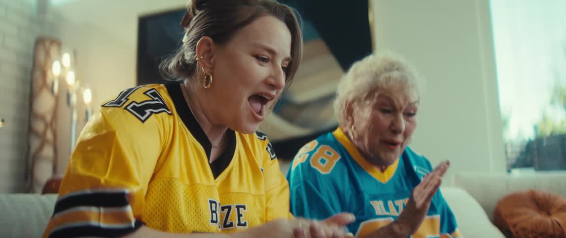 An excited grandmother and her granddaughter in opposing football jerseys celebrate wildly in slow motion while watching the big game at home