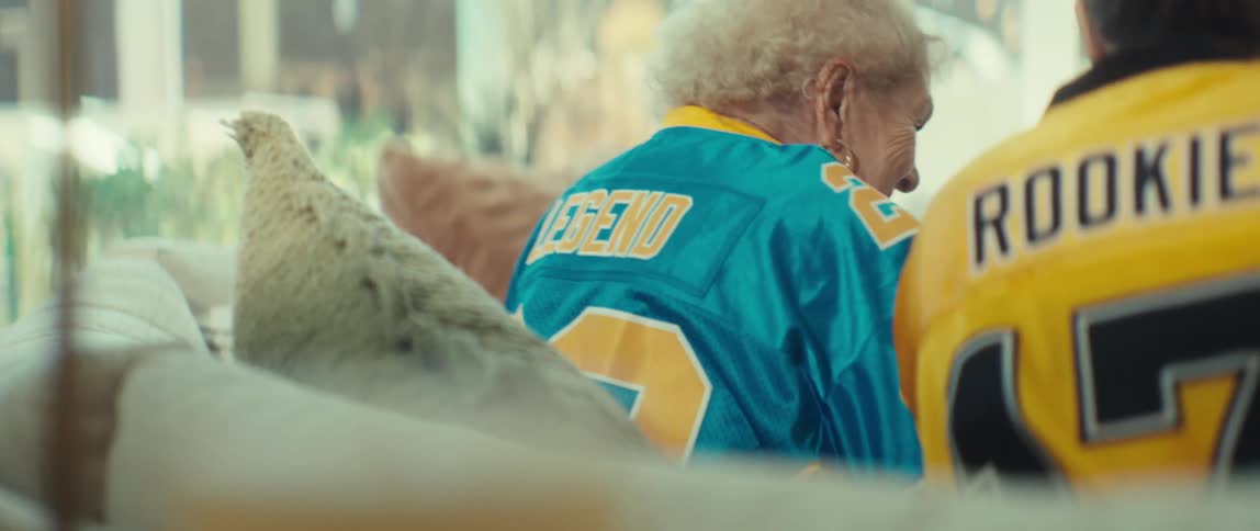 A slow pan reveals an elderly 'Legend' and a young 'Rookie' sitting side-by-side in football jerseys, cheering while watching a game
