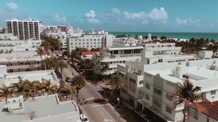 A slow aerial pull-out reveals the bustling streets and classic Art Deco architecture of Miami's South Beach, with the ocean visible in the background