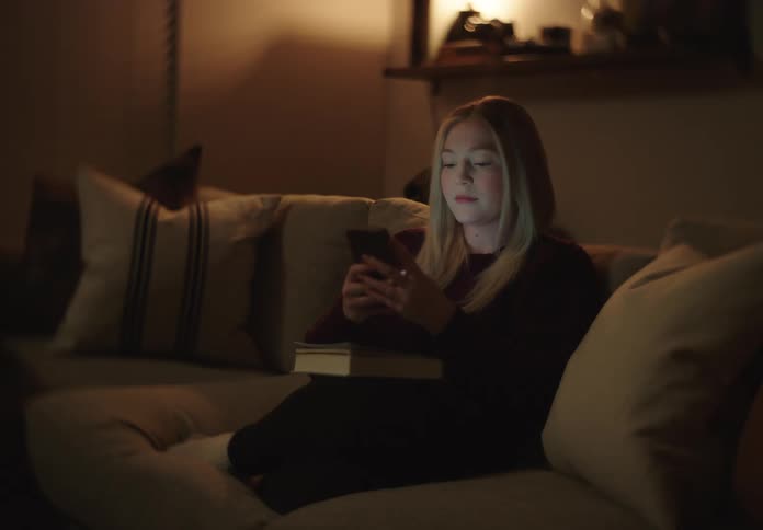 A young woman sits on a couch in a dimly lit living room, illuminated by the glow of her smartphone as she scrolls