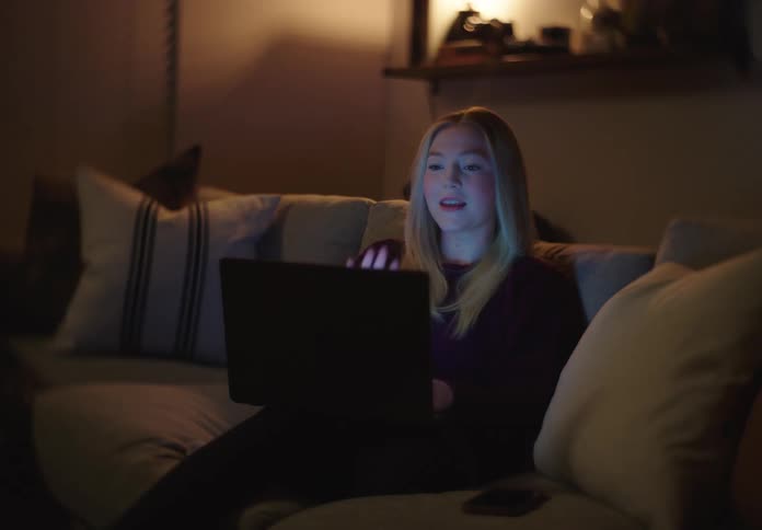 A young woman smiles and talks while on a video call on her laptop in a cozy, dark living room