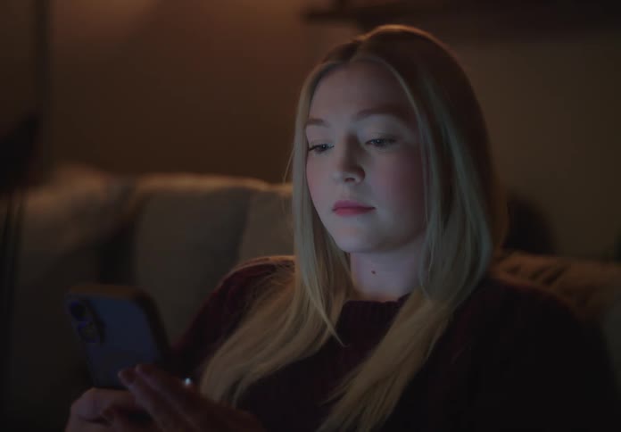 A young blonde woman scrolls on her smartphone at night in a dark room, her face illuminated by the screen's glow