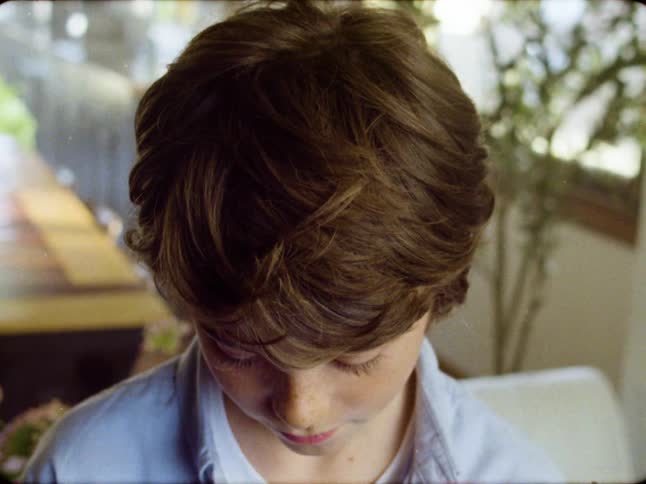 A nostalgic Super 8 style close-up shot captures a young boy with brown hair looking down in quiet contemplation