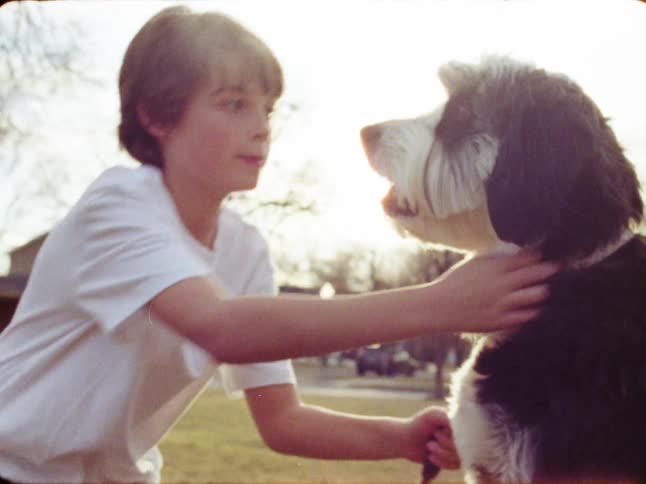 A nostalgic vintage home video clip shows a young boy lovingly petting and kissing his fluffy dog on a sunny day