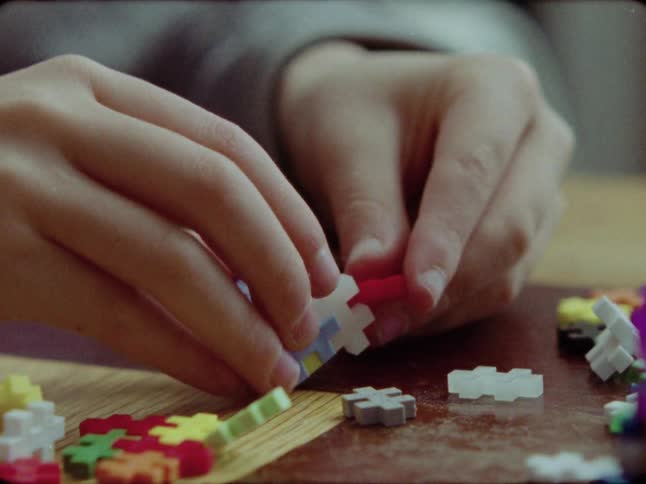 A close-up shot of a child's hands building a colorful object with small interlocking plastic puzzle pieces on a wooden table