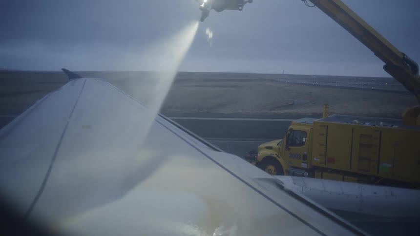 From a passenger's window view, a de-icing truck sprays an airplane's wing with fluid before takeoff on a cold day