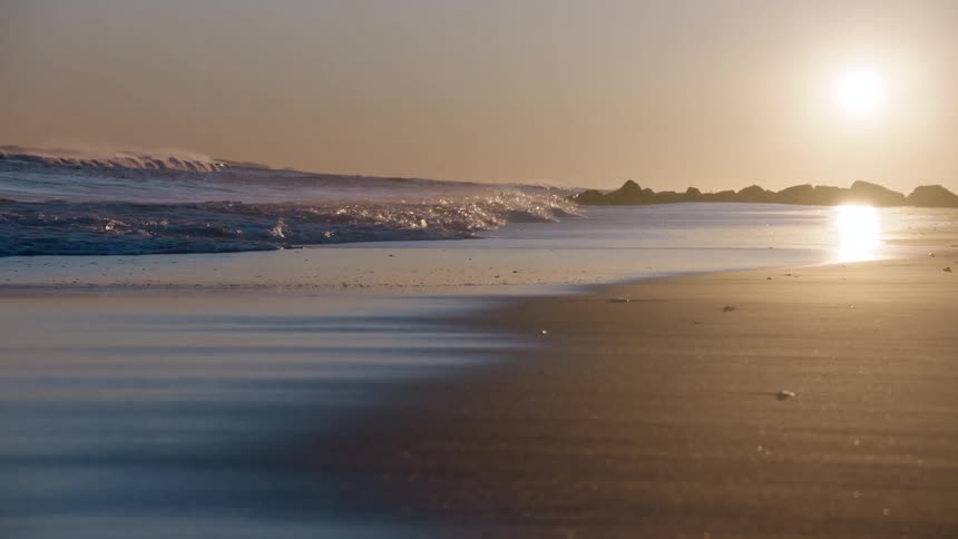 Ocean waves break in slow motion along a sandy beach as the golden sun sets beautifully over the water