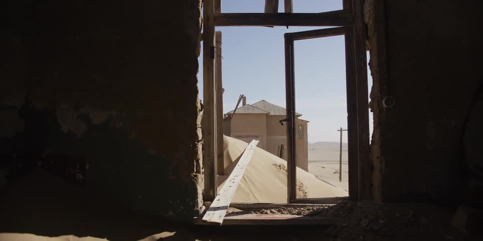 A slow zoom-out from a broken window reveals the sand-filled interior of an abandoned building in a desert ghost town