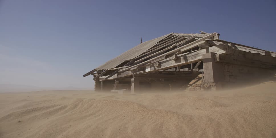 Strong winds desert sand across a dune, slowly engulfing a dilapidated and abandoned home