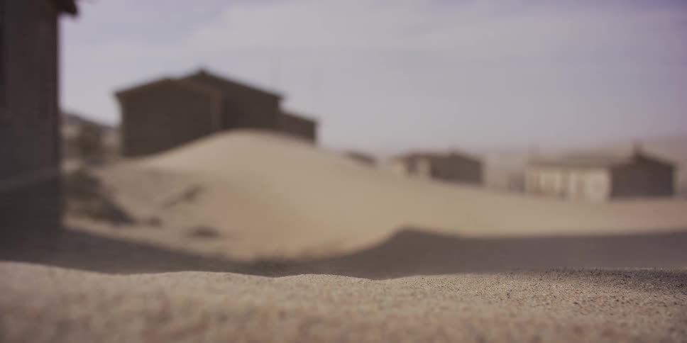 Wind sweeps sand across the ground in slow motion with a blurry, abandoned desert town in the background