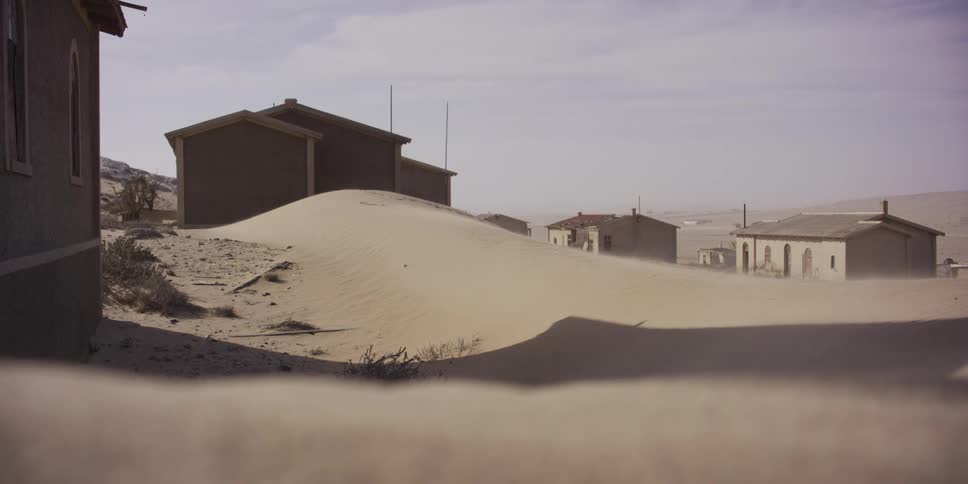 A static shot of an eerie abandoned ghost town in the desert with sand blowing over large dunes