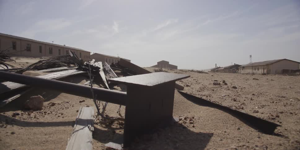 A slow tracking shot across the desolate, sandy ground of a deserted ghost town reveals weathered debris and crumbling buildings
