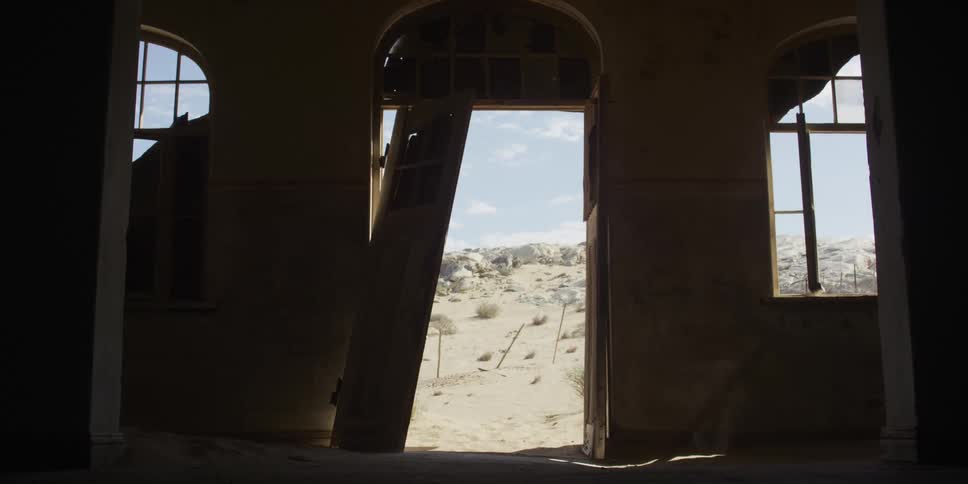 Wind blows sand through the broken doorway of a dark, abandoned building, revealing a bright desert landscape outside