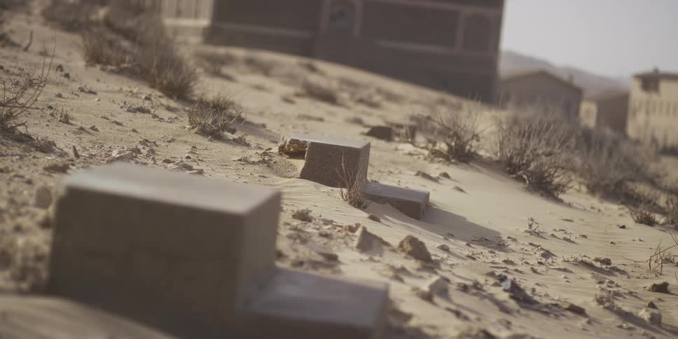 Close up on forgotten concrete steps buried in the desert sand as a sandstorm blows past abandoned buildings