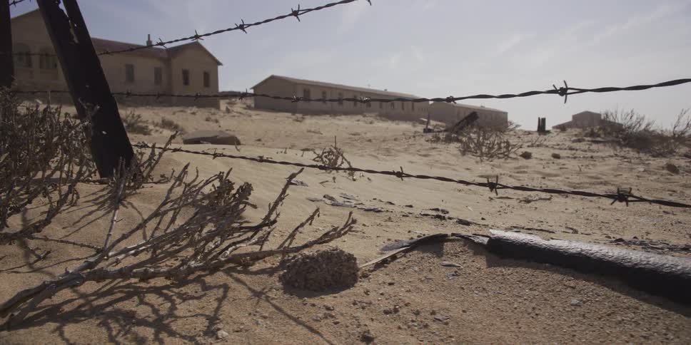 A slow, low-angle slider shot moves along a barbed-wire fence in a desolate, sand-swept ghost town