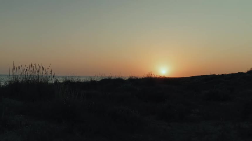 The sun sets over the ocean horizon, viewed from behind tranquil, grassy sand dunes