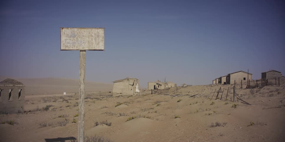 A weathered wooden sign stands in front of crumbling buildings in an abandoned desert ghost town