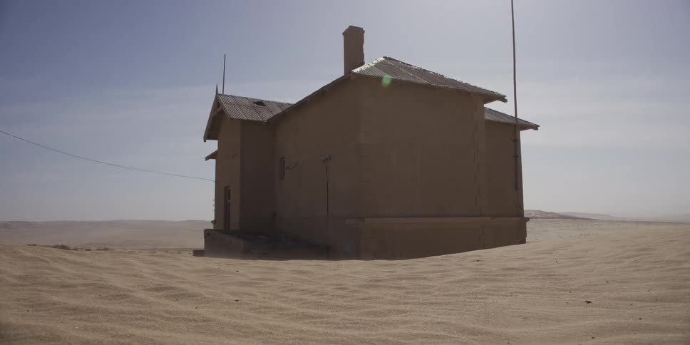 A desolate abandoned house is slowly being consumed by the shifting desert sands as wind blows across the arid landscape