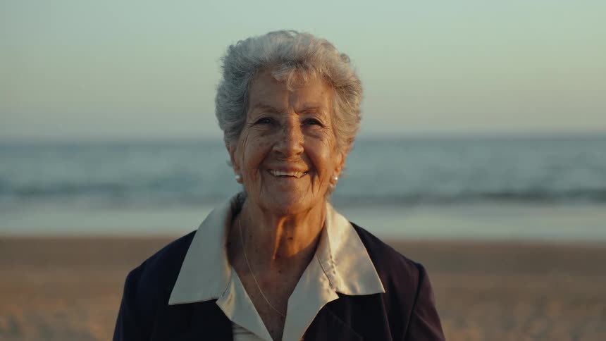 Close-up portrait of a serene and happy elderly woman smiling warmly at the camera on a sandy beach at sunset