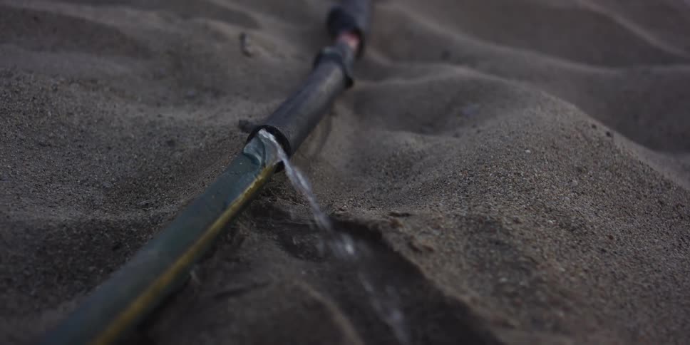 A damaged irrigation pipe leaks water into the arid sand in slow motion