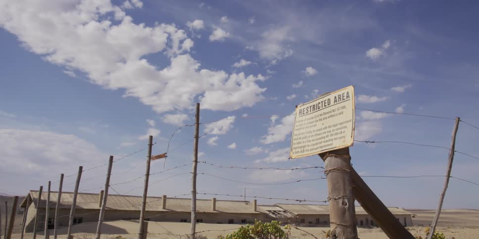 Zooming out from a 'Restricted Area' sign on a barbed-wire fence in front of a derelict desert compound under a cloudy blue sky