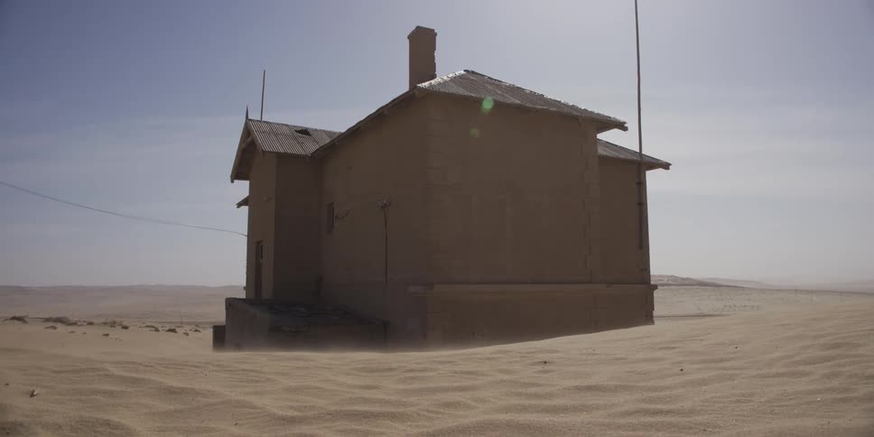 A desolate, abandoned house stands alone in the desert as strong winds sand across the landscape, slowly burying the structure