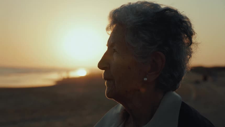 A serene elderly woman watches the golden sunset from the beach, a gentle smile on her face