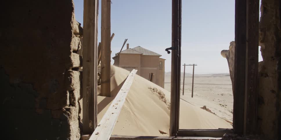 Dolly shot through the doorway of a ruined desert home, revealing a ghost town slowly being buried by sand dunes