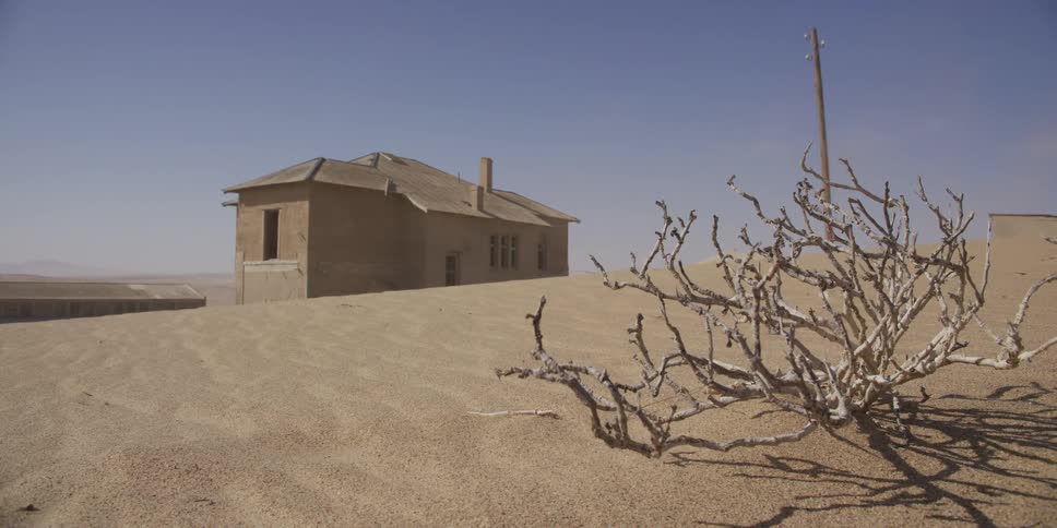 Wind blows sand across an abandoned building partially buried in the dunes of a desert ghost town