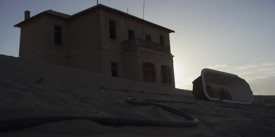 A low-angle view shows an abandoned house and bathtub partially buried in the desert sand at dusk