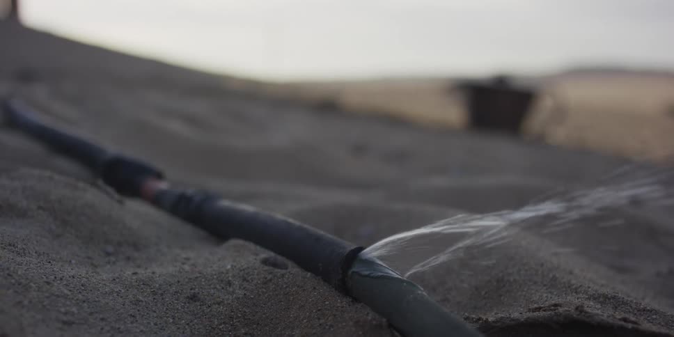 Slow motion close-up of a cracked irrigation pipe spraying precious water onto the dry desert sand