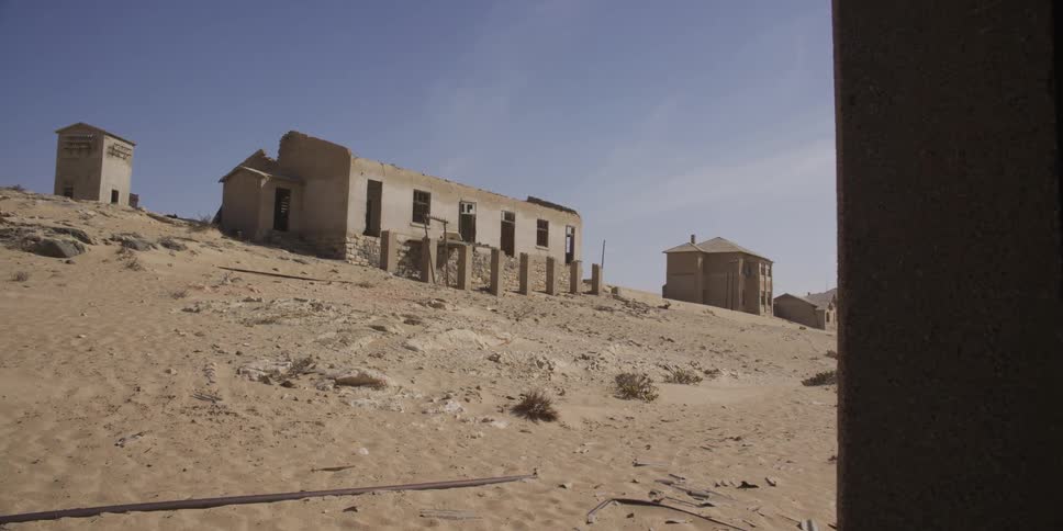 Pan shot reveals the desolate, sun-scorched landscape and ruined buildings of an abandoned desert ghost town