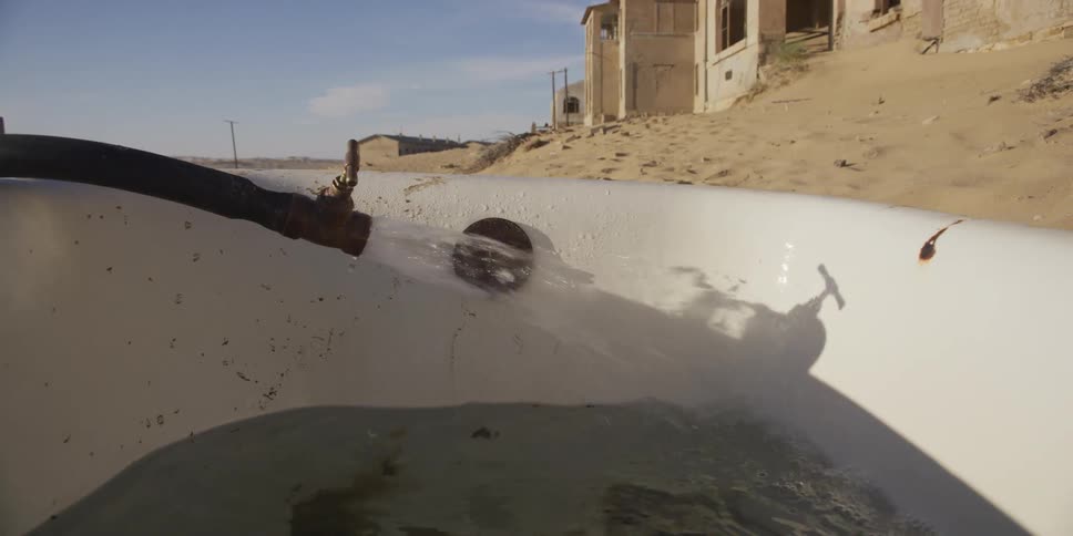 Water pours from a hose into an old bathtub sitting in the sand of an abandoned desert town