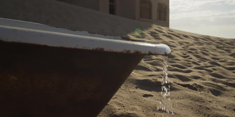 Water pours from a rusty bathtub in slow motion, wasted into the sand of a desolate, abandoned desert ghost town