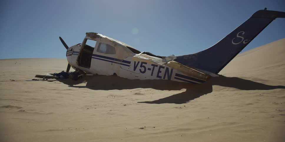 A wrecked propeller plane lies half-buried in the sand of a vast desert under a clear, bright sky