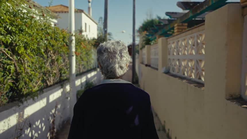 Slow motion shot from behind of an elderly woman with grey hair walking down a sunny residential alley